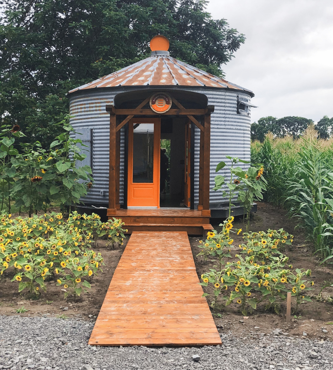 Enright Cattle Company. A tin silo shaped shed with a ramp and orange door. Flowers surrounding it.